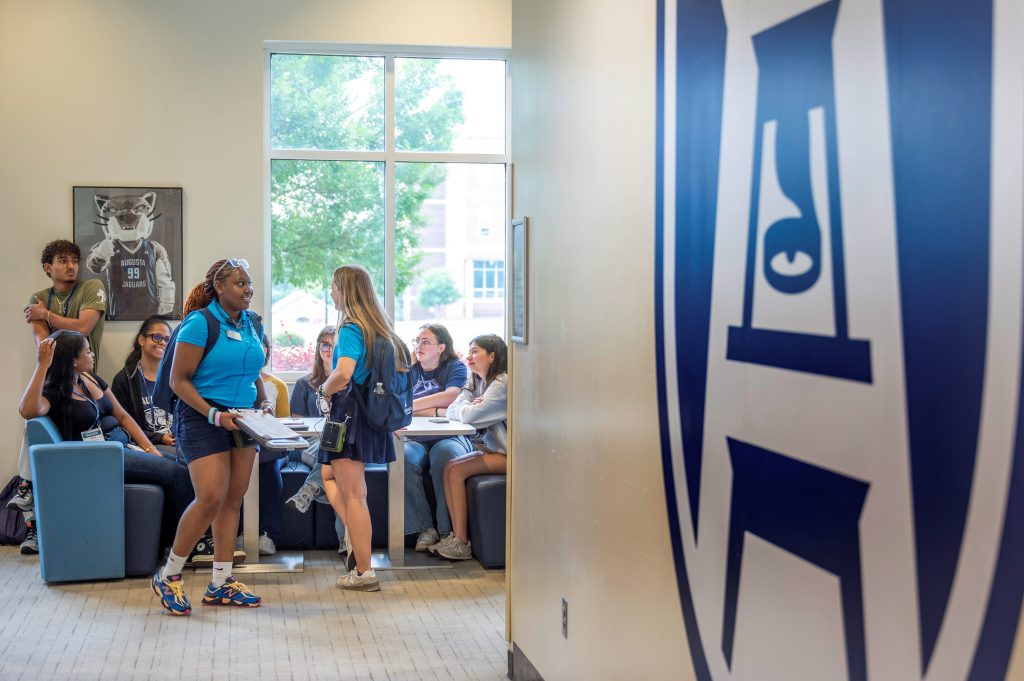 A group of men and women, some standing and some are sitting at a table, talking to one another.