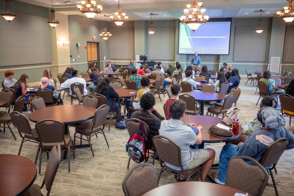 Men and women seated at round tables in a large room listen attentively to a man speaking at the front of the space.