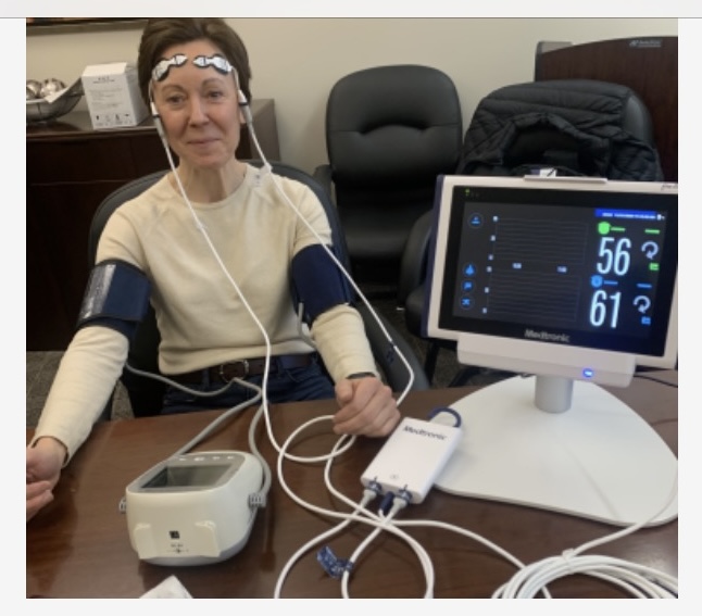 A woman smiles at the camera, she's hooked up to a monitor with a lot of cords and sensors on her head and around her arms