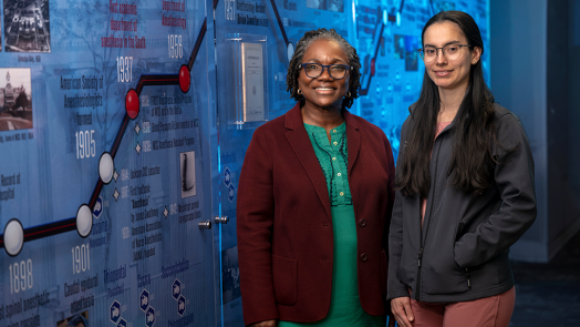 Two female anesthesiologists pose in front of a background with the history of MCG anesthesiology on it.
