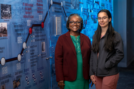 Two female anesthesiologists pose in front of a background with the history of MCG anesthesiology on it.