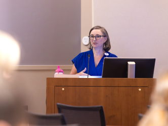 A woman presents to a large crowd of medical students and doctors in a large classroom.