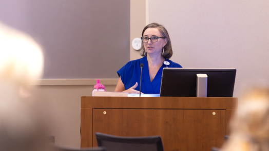 A woman presents to a large crowd of medical students and doctors in a large classroom.
