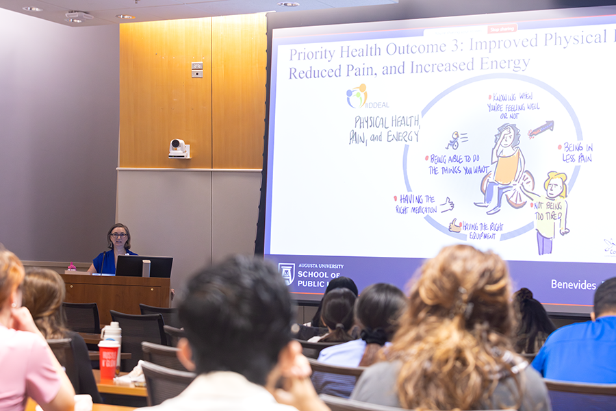 A woman presents to a large crowd of medical students and doctors in a large classroom.
