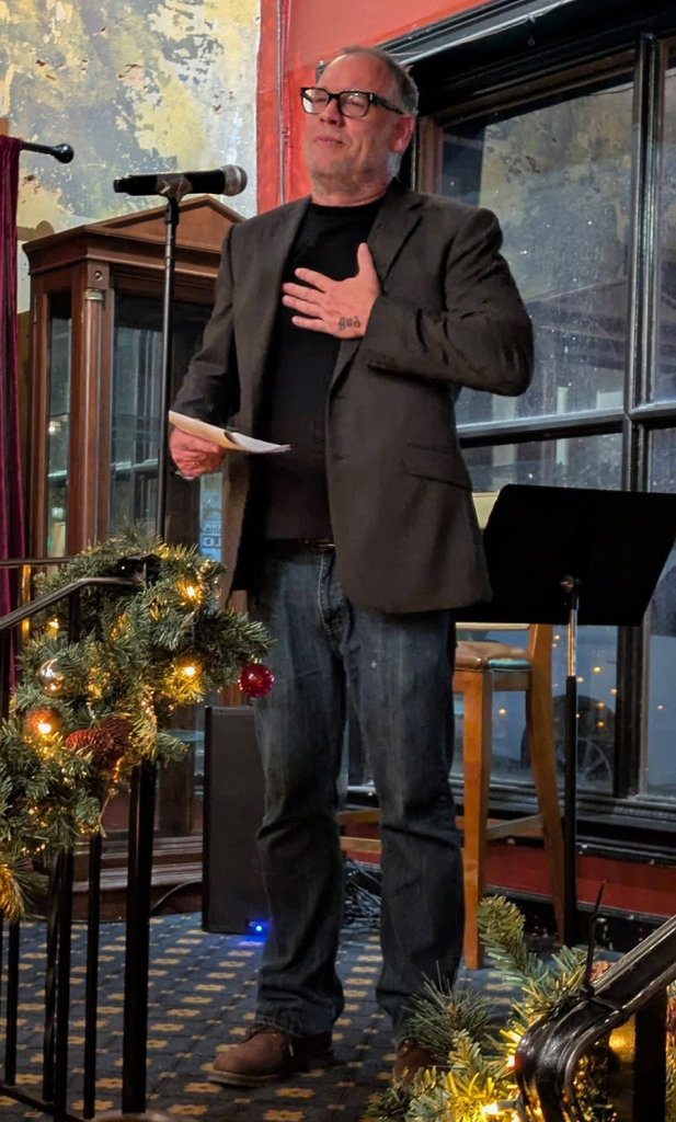 A man standing in front of a group of people in a room and speaking to them around Christmas time.