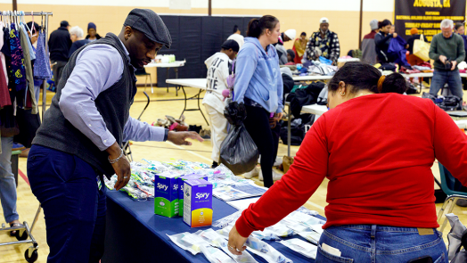 A man stands behind a table with toiletry items on it while a woman picks something up from it. People in the background do them same.