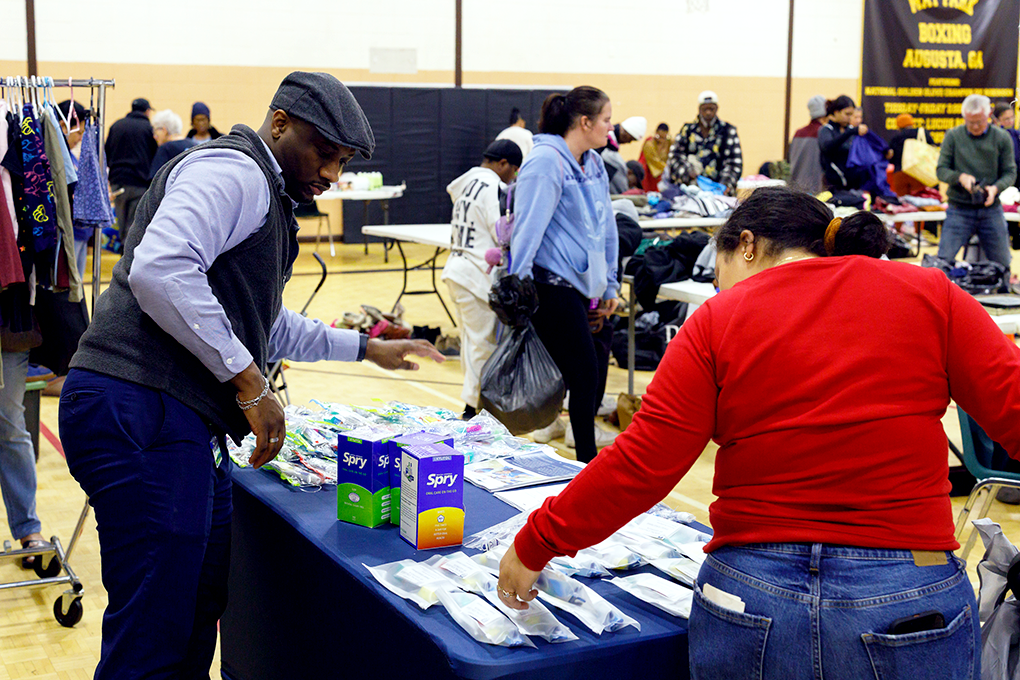 A man stands behind a table with toiletry items on it while a woman picks something up from it. People in the background do them same.