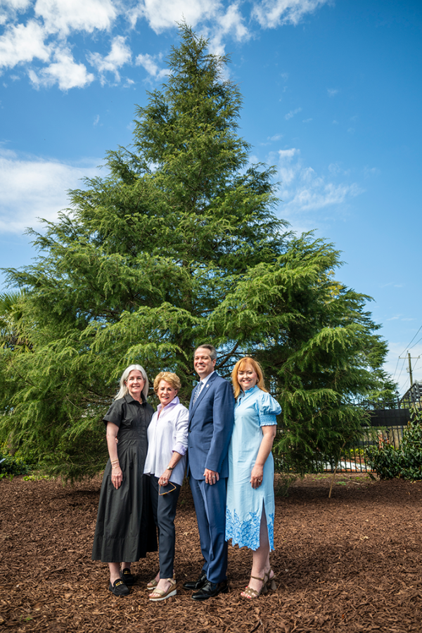 Three women and one man stand in front of a large tree.