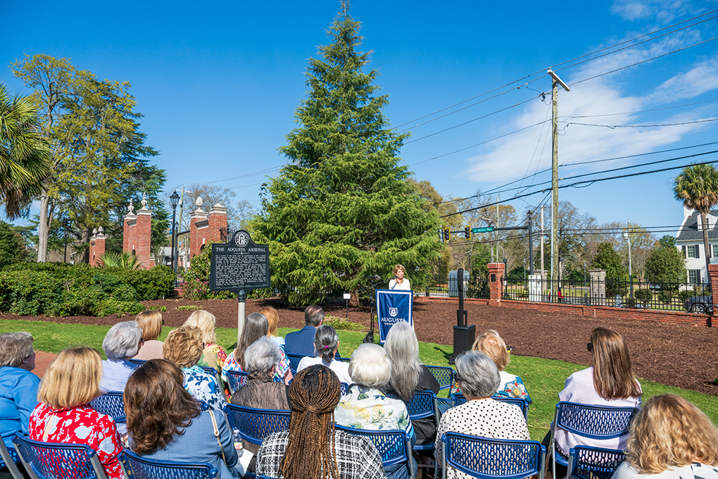 A woman at a podium and three rows of women in chairs.