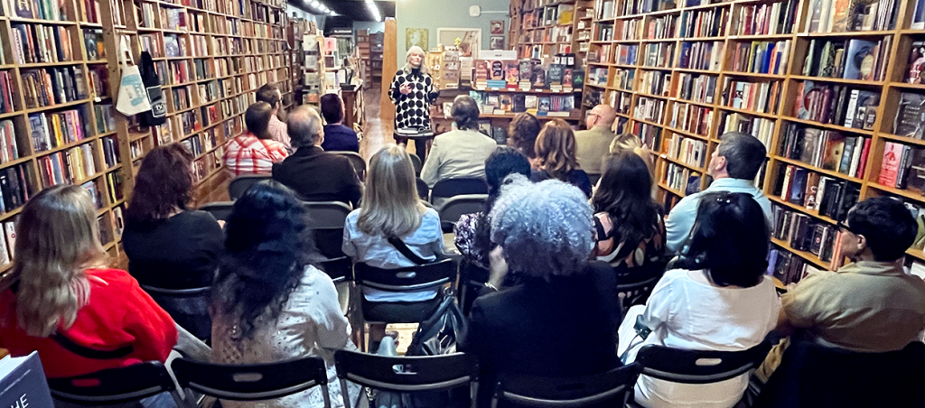 A woman stands in a library in front of a group of people and speaks to them.
