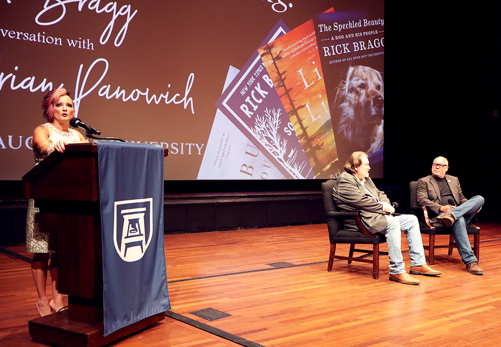 Two men sit in chairs on a stage while a woman stands behind a podium and speaks to an audience.