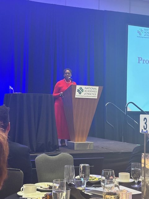 A woman in formal attire stands on a stage, speaking into a microphone behind the podium