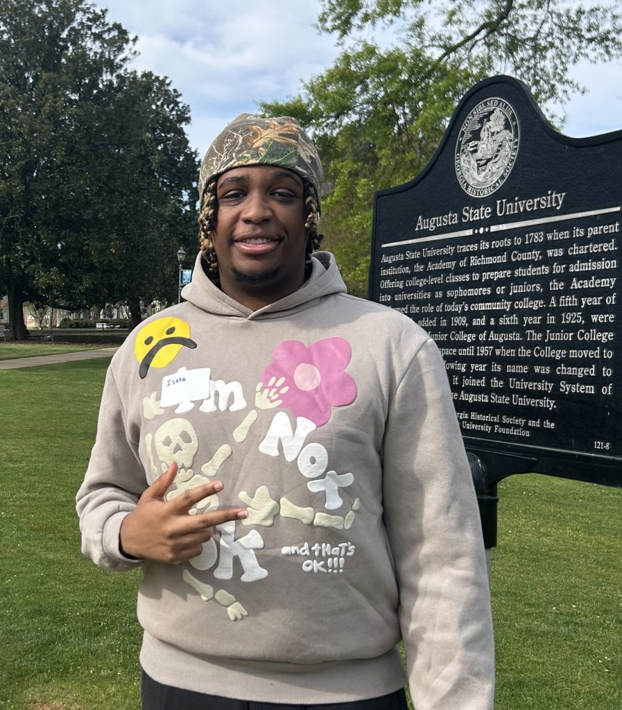 A man wearing a beanie and sweatshirt poses for a photo while standing in front of a large metal plaque. 