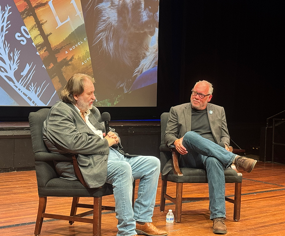 Two men sit in chairs on a stage and talk to each other with micophones.