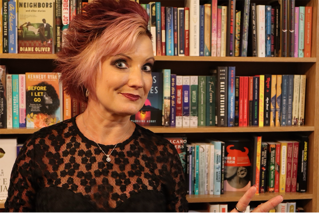 A woman stands in front of a book shelf and smiles at the camera.