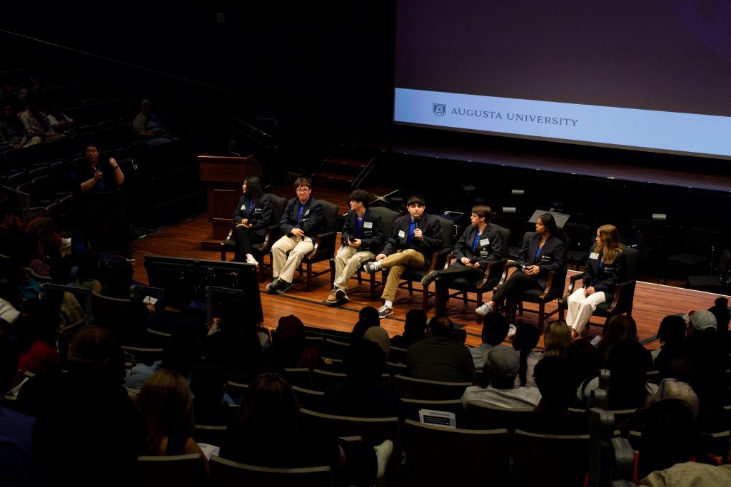 A group of men and women sit in a single file line of chairs on top of a stage.