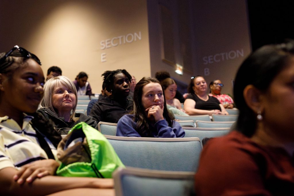 Several men and woman, looking ahead, sit inside of a theatre.
