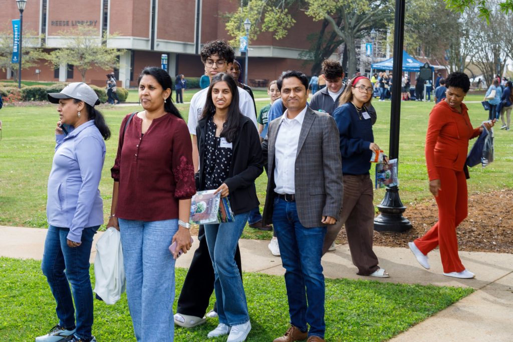 Three women and one man pose for a photo outdoors as a crowd passes behind them.