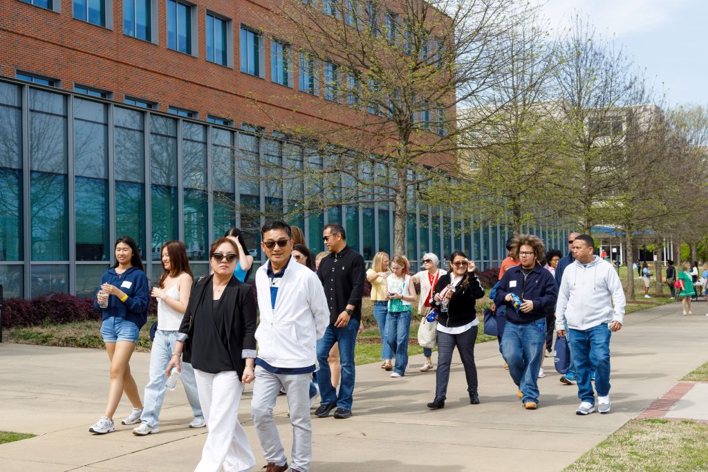 A large group of men and woman walk down the sidewalk with a large building in the background. 