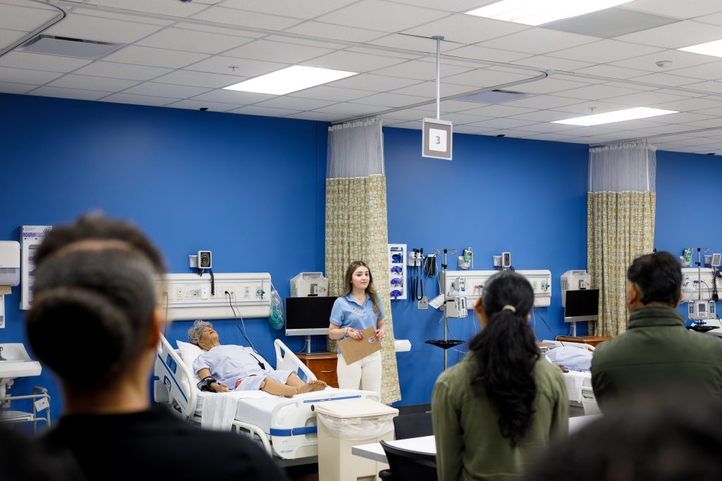 A woman stands near a medical dummy while speaking to a large crowd.