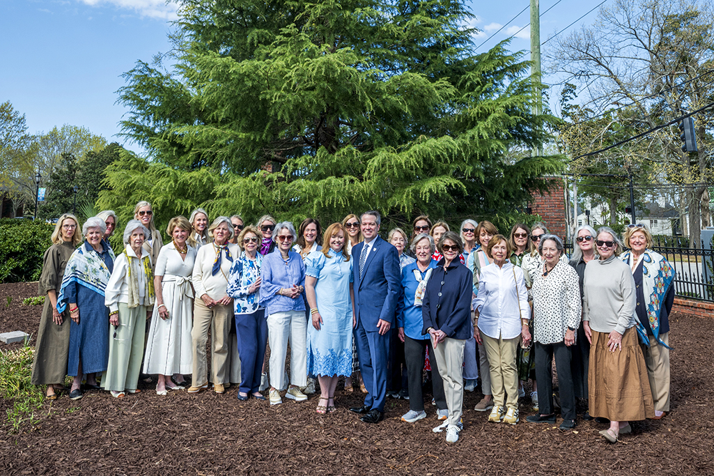 Large group of ladies and one man in front of a large tree.