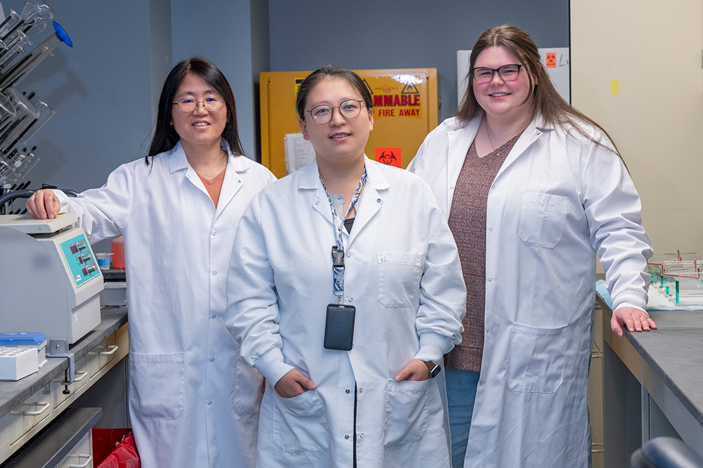 Three female scientists wearing lab coats smile at the camera.