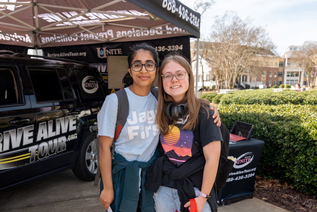 Two female students stand arm in arm, smiling for a photo in front of the impaired driving simulation vehicle.