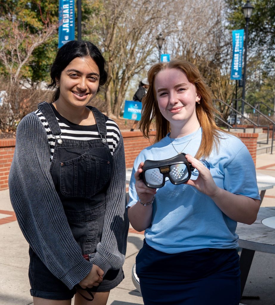 Two girls pose for a photo, with one holding up a pair of drunk goggles. 