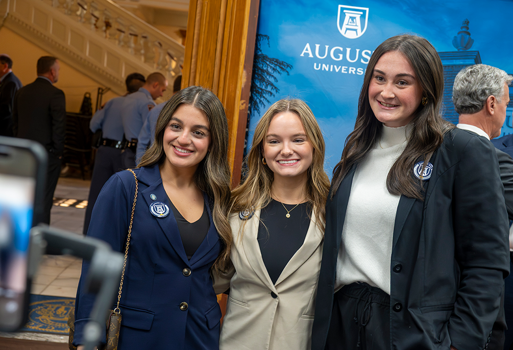 Three young women in business dress smile with blue screen behind them.