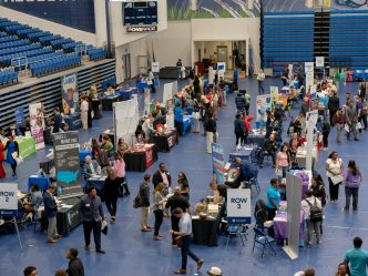 A large group of people stand talking at various indoor tabling opportunities.