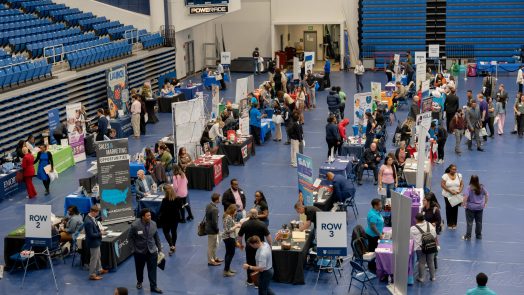 A large group of people stand talking at various indoor tabling opportunities.