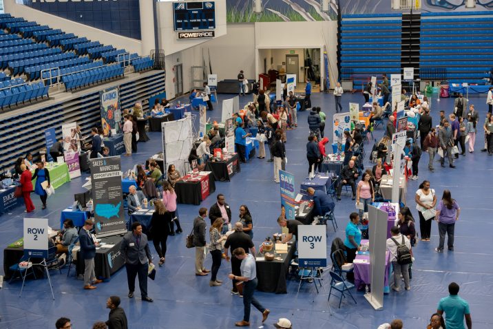 A large group of people stand talking at various indoor tabling opportunities.