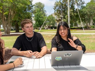 College-aged men and women sit at an outdoor picnic table near a computer.