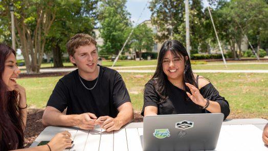 College-aged men and women sit at an outdoor picnic table near a computer.