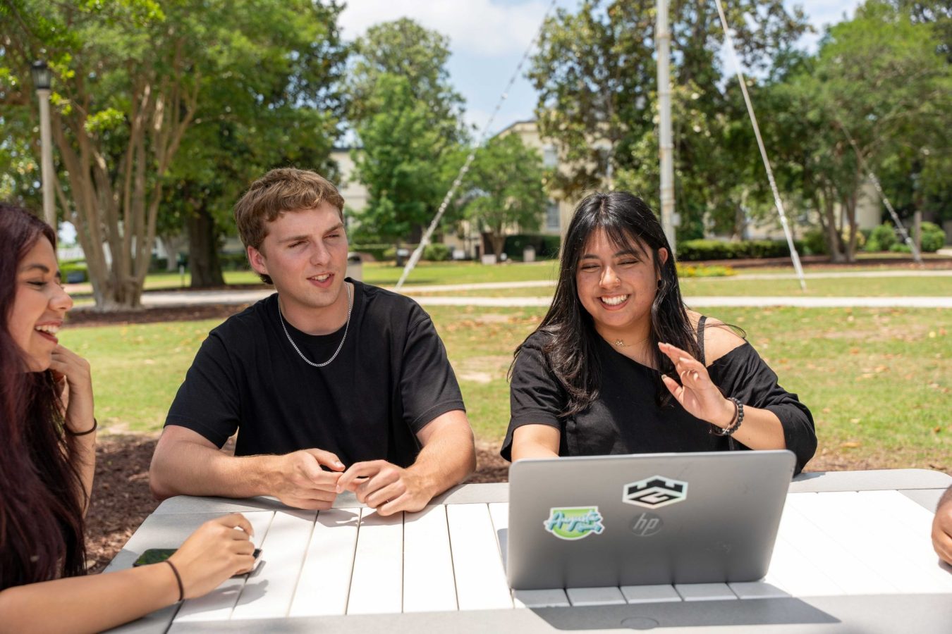 College-aged men and women sit at an outdoor picnic table near a computer.