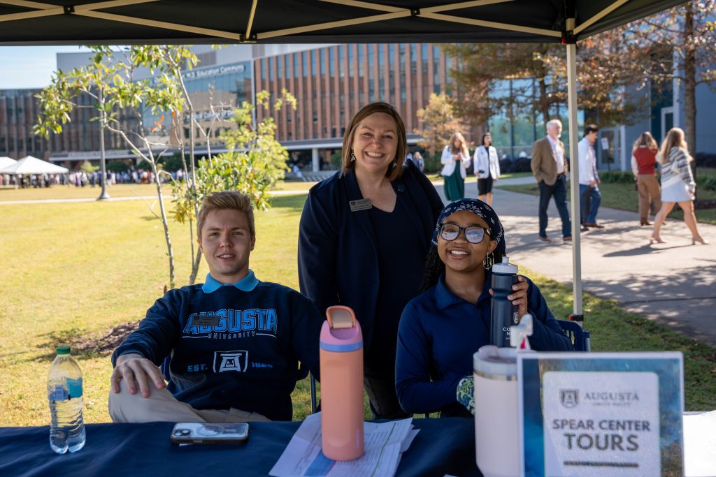 Two women and one man sit under a tent while tabling outside.