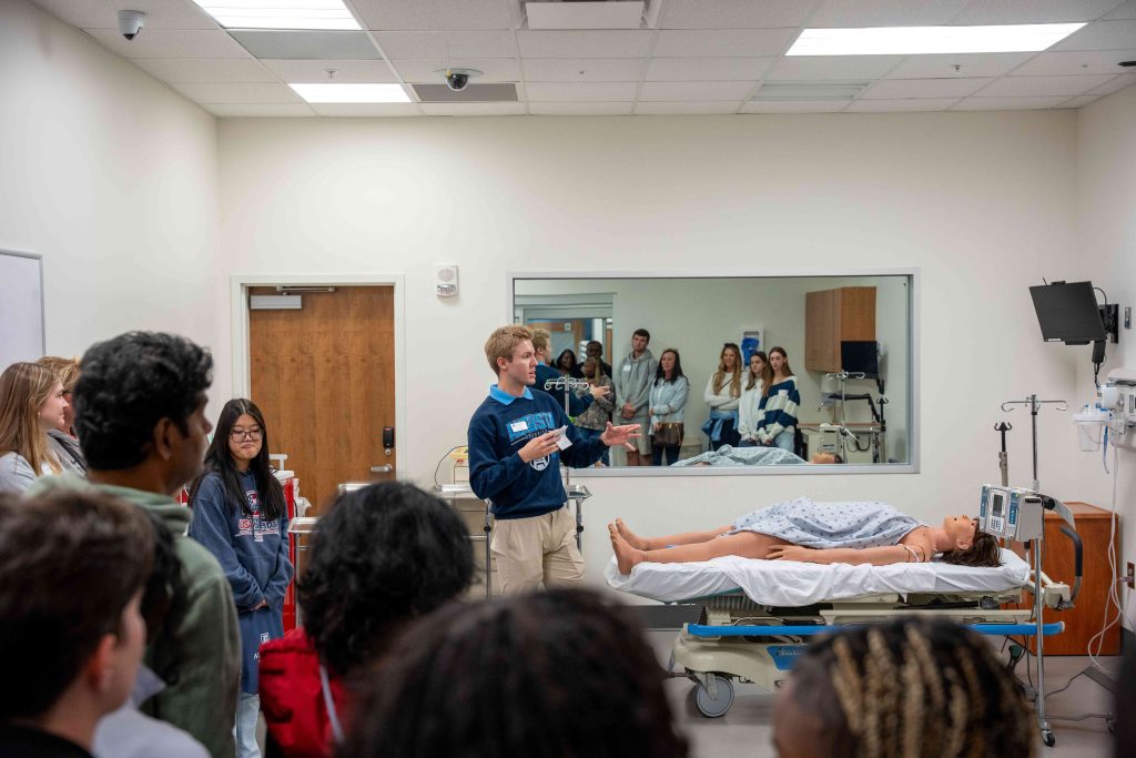 A large group of people surround a young man who is speaking over a medical dummy. 