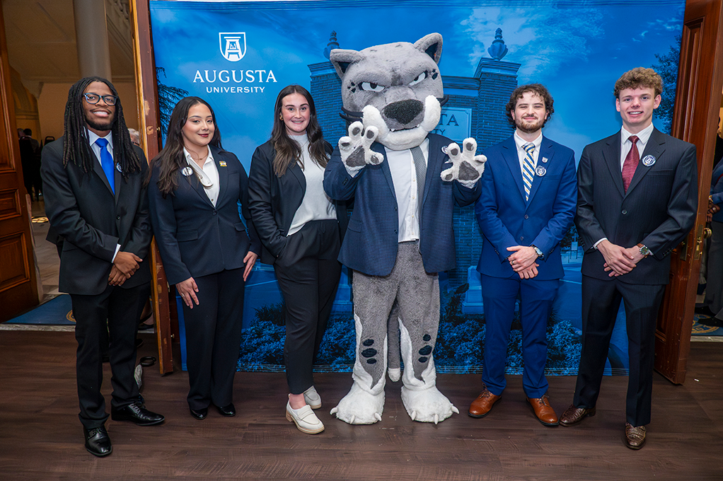 Three young men and two young women in business dress smile in a line with a costumed cat.
