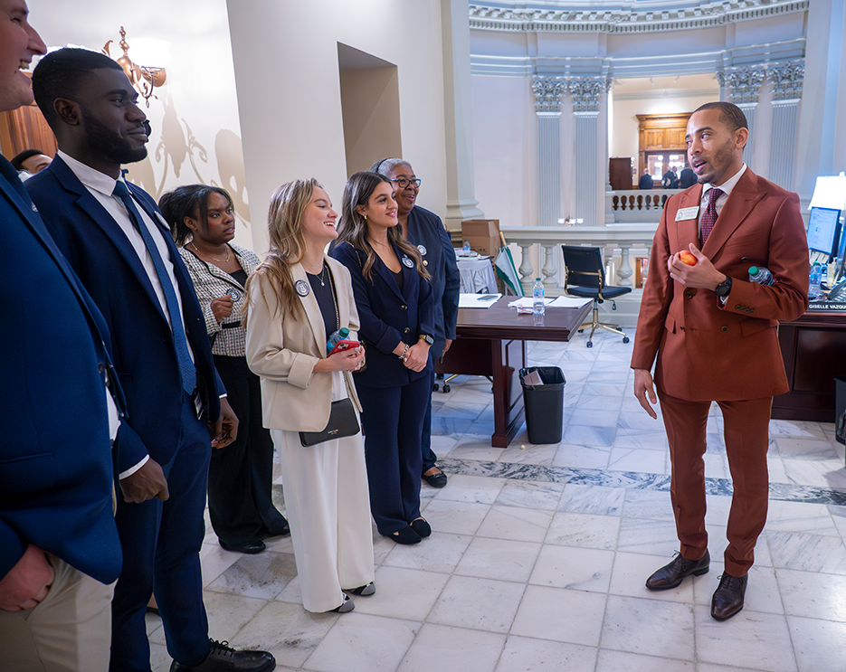Young men and women in various business dress listen to a man talking to them holding a snack.
