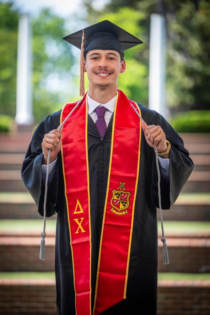 A man wearing a graduation cap and gown, while holding up his hands, smiles for a photo. 