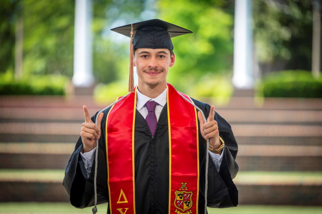 A man wearing a graduation cap and gown, while holding up his hands, smiles for a photo. 