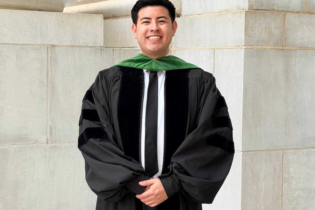A man wearing a graduation gown and a tie smiles at the camera.