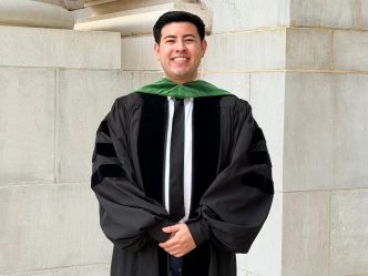 A man wearing a graduation gown and a tie smiles at the camera.