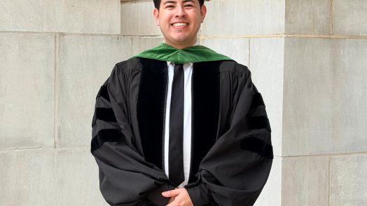 A man wearing a graduation gown and a tie smiles at the camera.