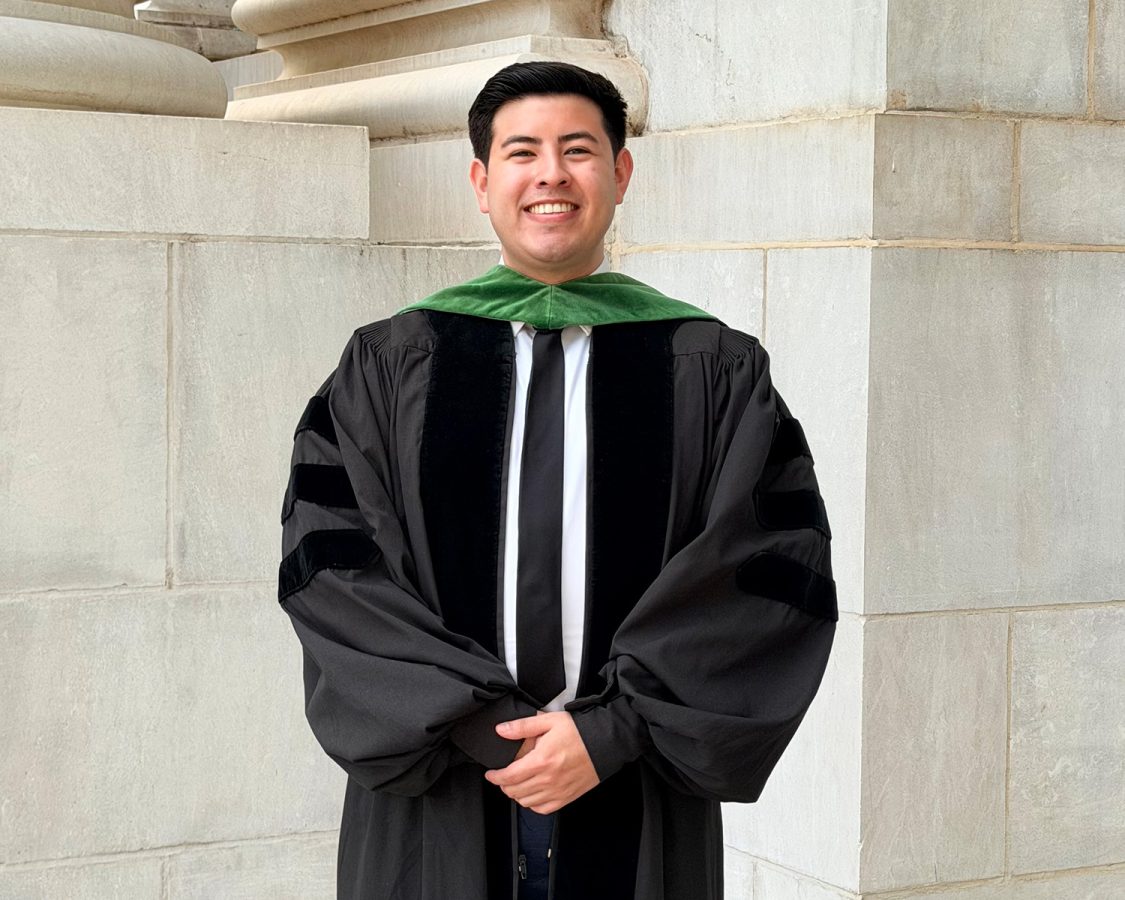 A man wearing a graduation gown and a tie smiles at the camera.
