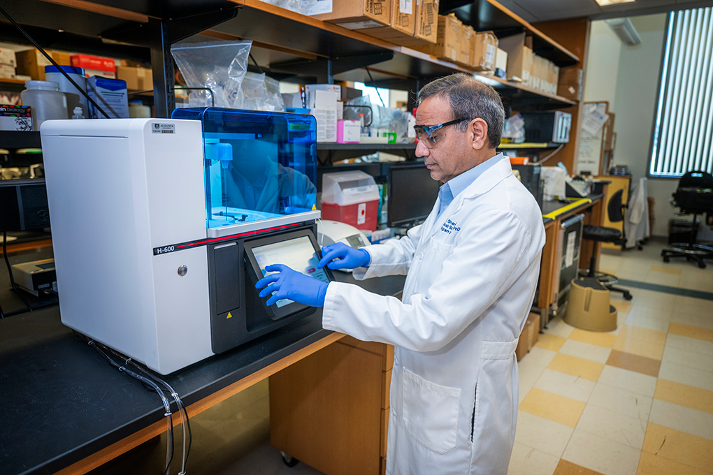 A male scientist wearing a lab coat, gloves, and goggles looks at a screen on a machine in a lab.