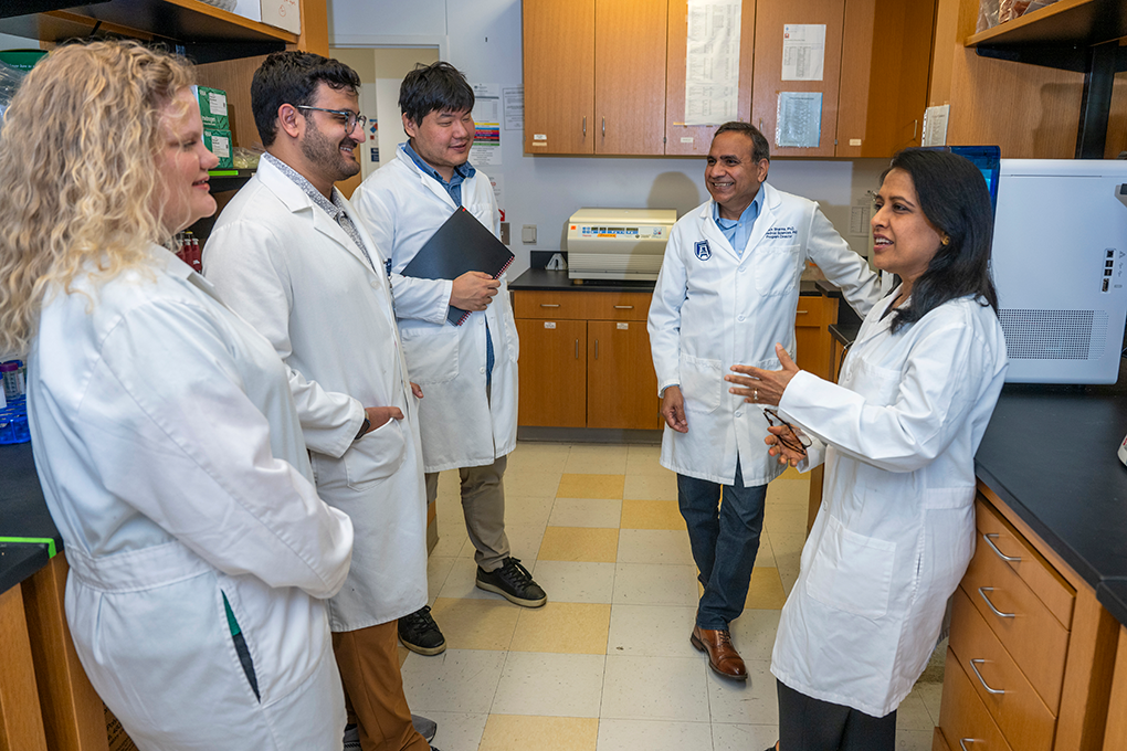 A group of scientists wearing lab coats stand around a machine in a lab and talk.