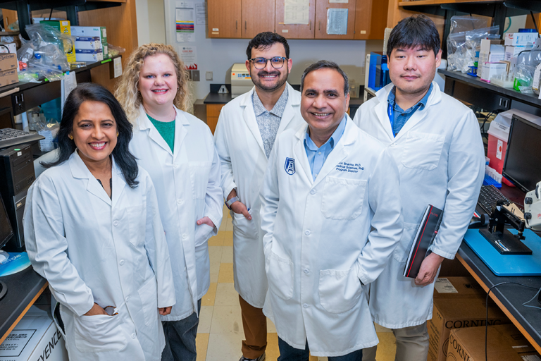 A group of male and female scientists wearing lab coats smile at the camera in a lab.