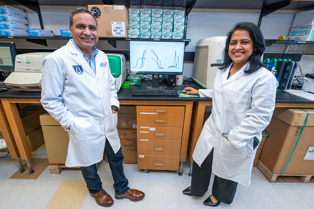 A man and woman scientist wearing lab coats stand in front of a computer in a lab and smile at the camera. 