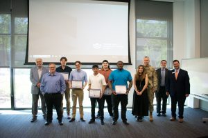 A group of men and one woman stand in front of a screen and hold awards.